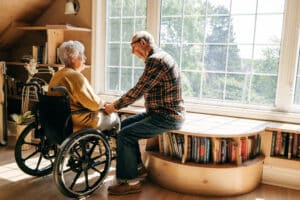 Ein altes Ehepaar reden miteinander vor einem großem Fenster in ihrem Haus. Die Frau sitzt im Rollstuhl